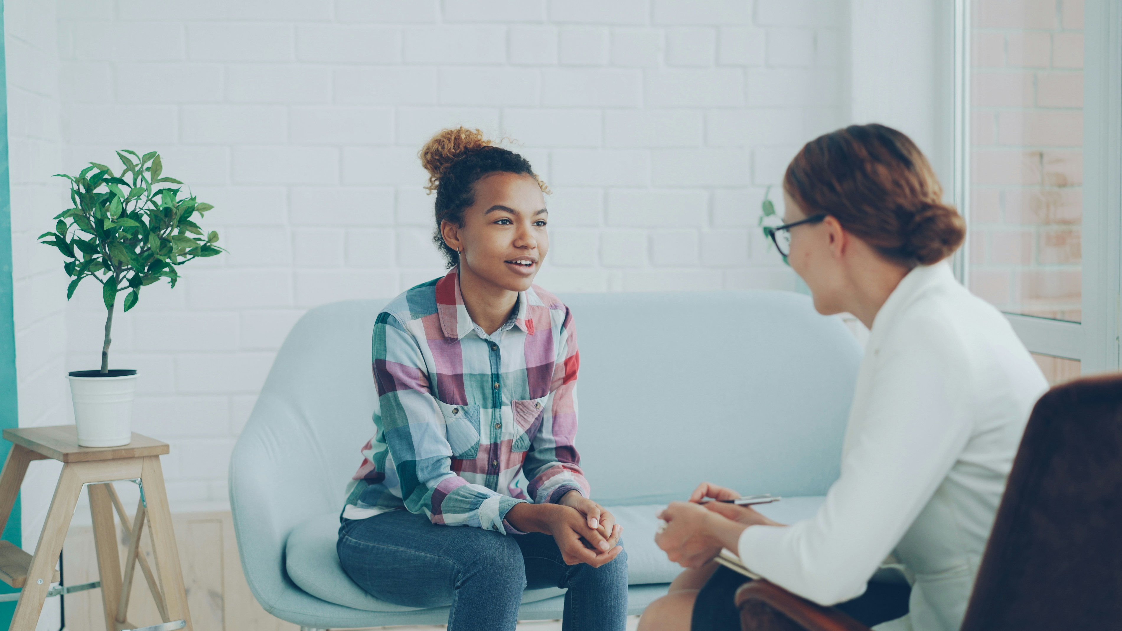 FULL 4K VIDEO for this still is in my bio link! ⬇️ Cheerful African American girl is talking to psychologist sitting on sofa in modern loft style studio and smiling while specialist is listening to her and making notes.